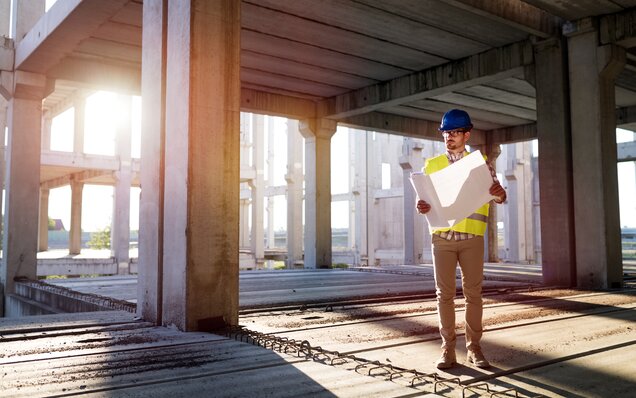 Civil engineer with helmet and safety vest checks construction site plans in a multi-storey rough concrete building at sunset